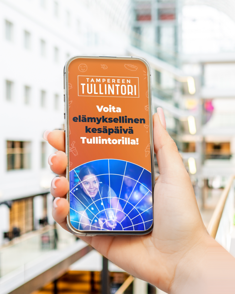 Kuvassa näkyy älypuhelin henkilön kädessä Tullintorin kauppakeskuksessa. Puhelimen näytöllä on Tullintorin kesäkampanjan 2026 raikas oranssi mainos, jossa lukee "Voita elämyksellinen kesäpäivä Tullintorilla!".